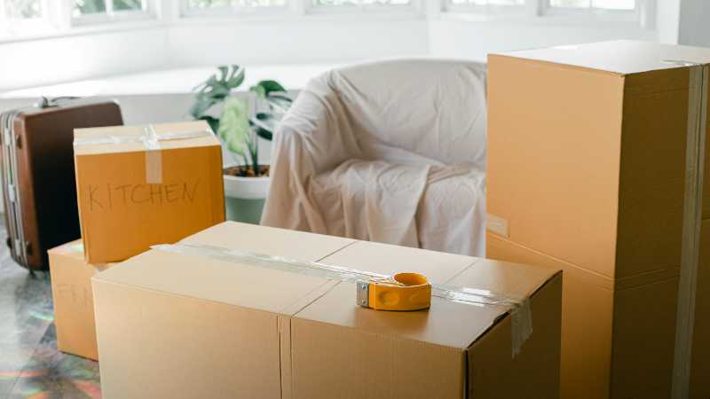 Cardboard boxes labeled 'Kitchen' in sunlit living room, ready for relocation., tags: packing - pexel